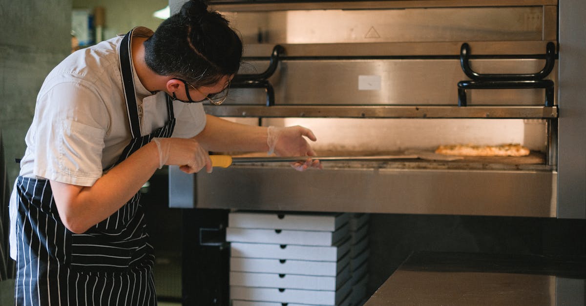 A chef in an apron prepares pizza in a commercial kitchen oven.